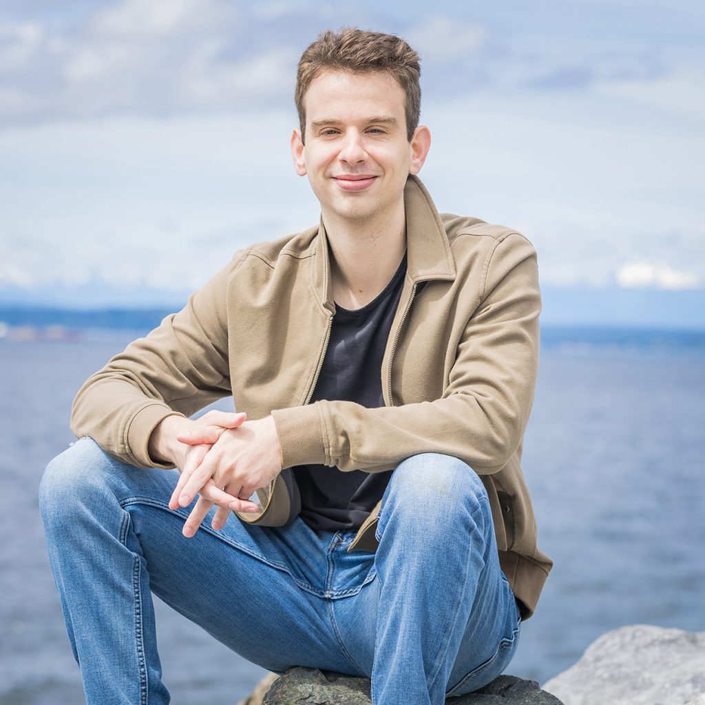 portrait of a young man sitting on a rock with ocean and mountain views behind him.